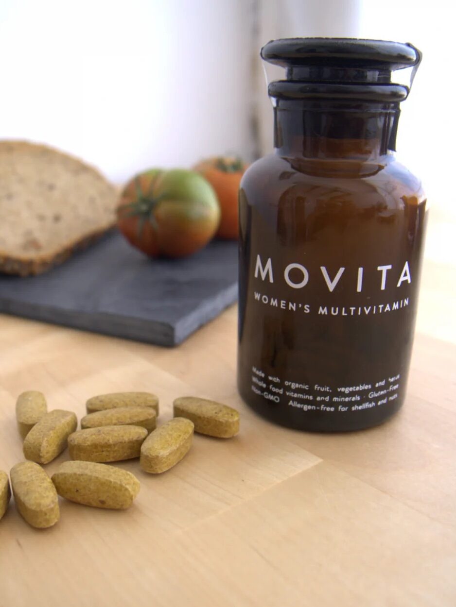 A bottle of movita women's multivitamins with pills scattered in front, a tomato and a slice of bread in the background on a wooden table.