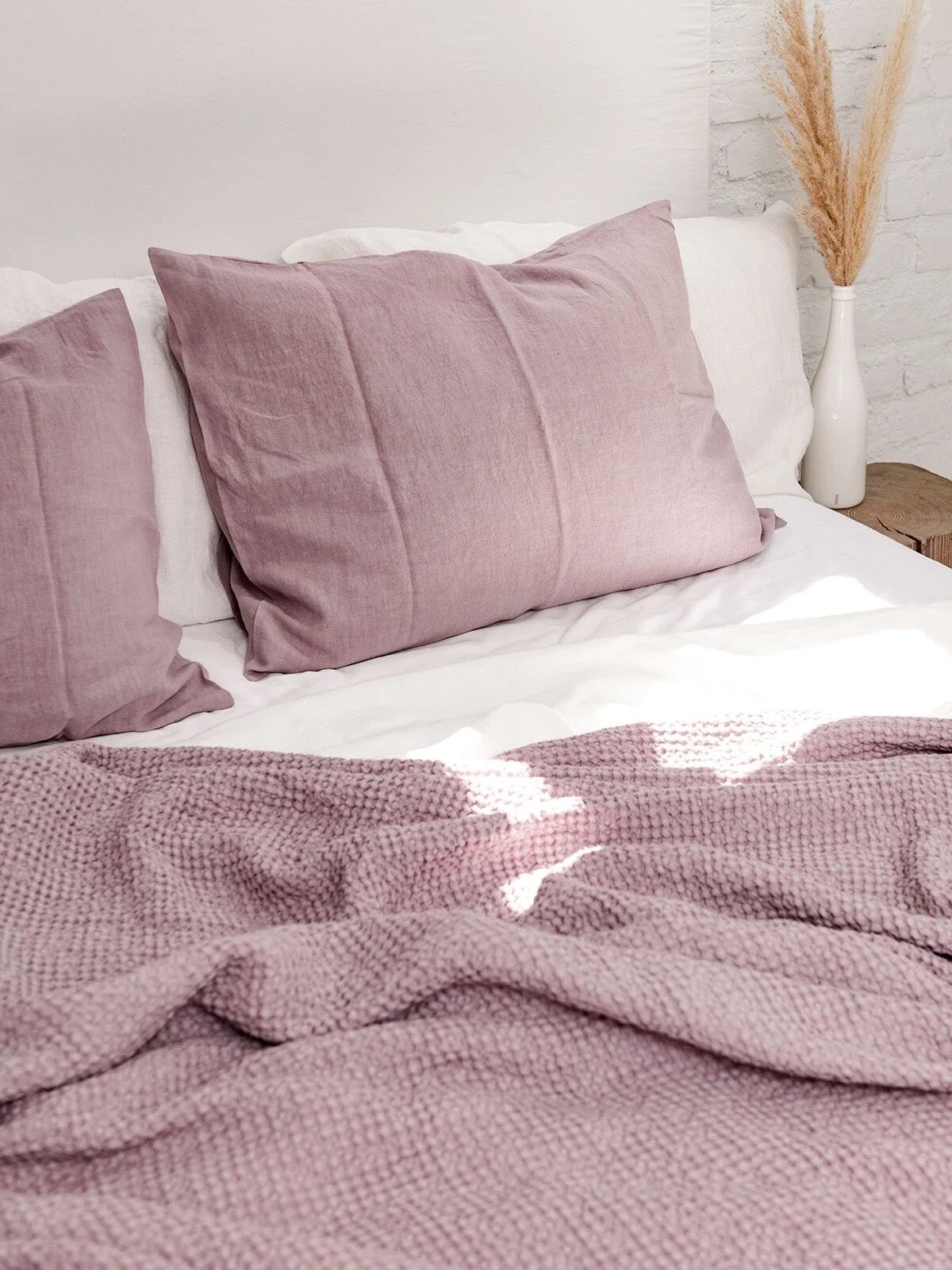 A neatly made bed with plush pink pillows and a textured pink throw blanket, set against a white wall with decorative dried plants.