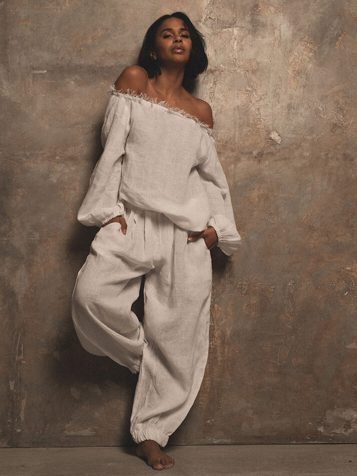 A woman in a casual off-shoulder white outfit stands against a textured beige background, posing with one leg bent and a serene expression.