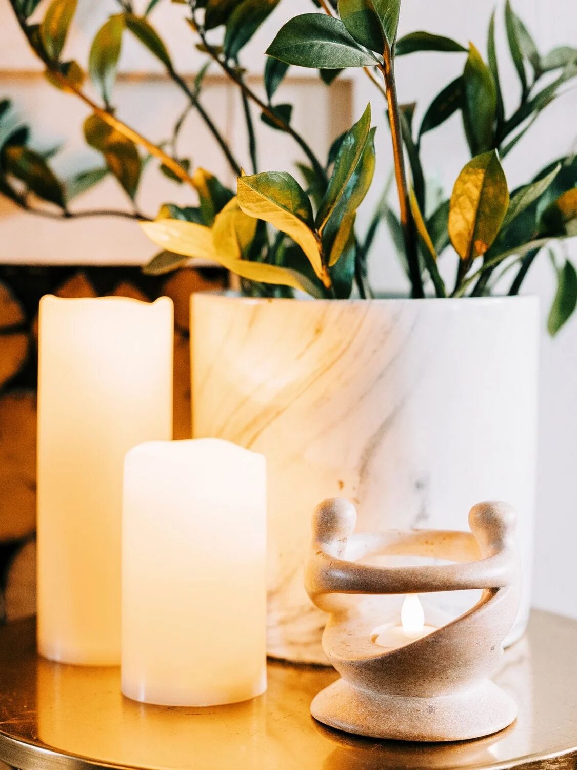 Two led candles and a small ceramic candle holder with a lit candle on a golden table beside a plant in a white pot.