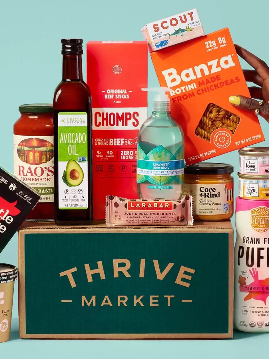 A person's hand holding a variety of packaged food products emerging from a "thrive market" box against a teal background.