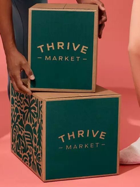 Two people holding thrive market boxes against a pink background, one wearing slippers and the other in socks.