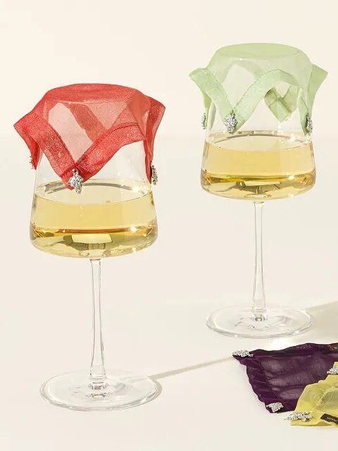 Three wine glasses with white wine, each covered by a different colored decorative mesh topper, arranged on a light surface with shadows.