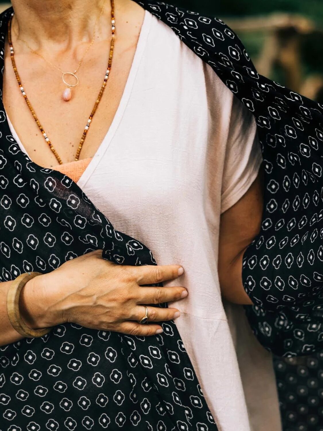 Close-up of a woman wearing a white top and black patterned shawl, her hand resting on her chest with visible necklaces and a bracelet.