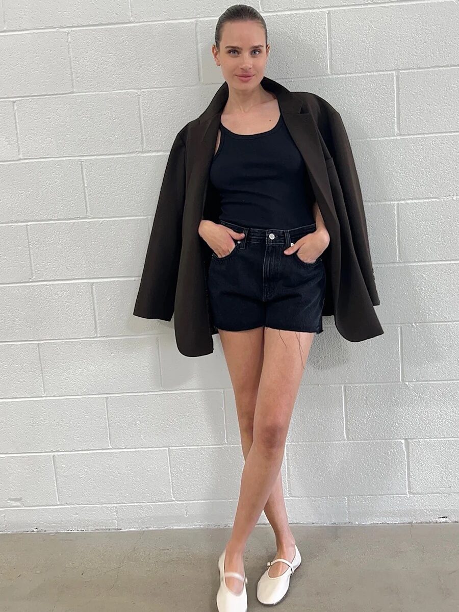 A woman standing against a white brick wall, wearing black shorts, a black top, a brown jacket, and white loafers.