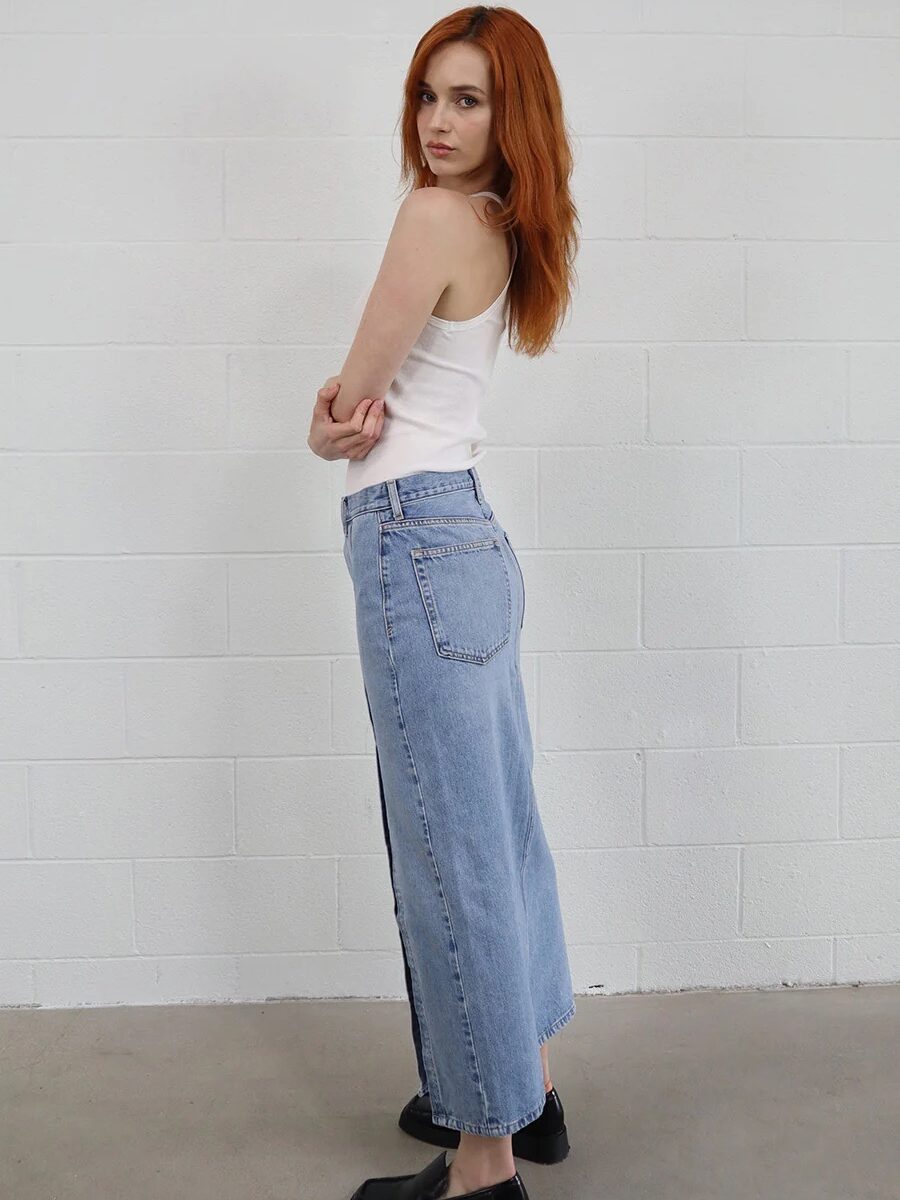 A woman with red hair wearing a white tank top and blue denim jeans stands against a white brick wall, looking over her shoulder.