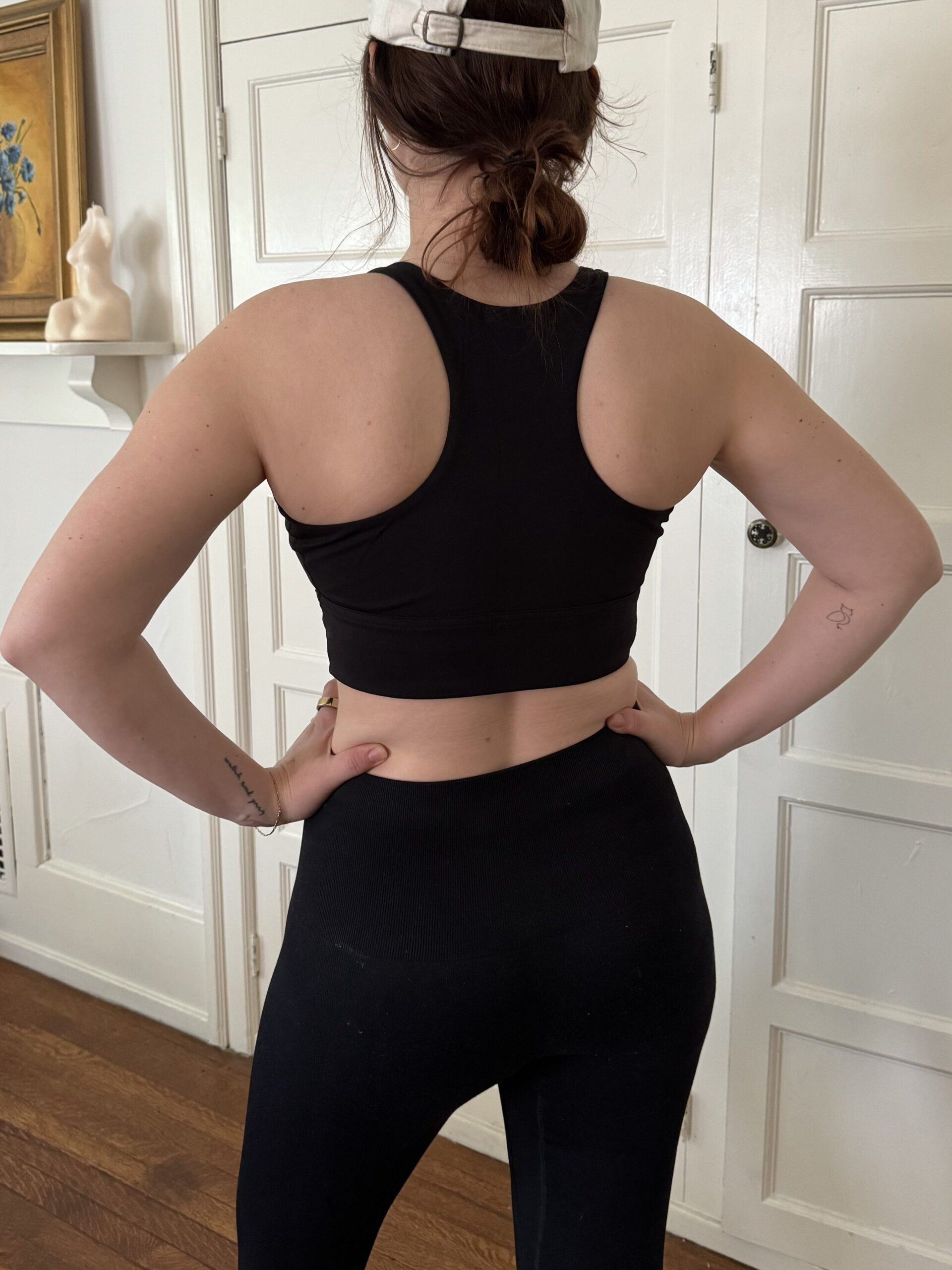 Woman wearing black sports bra and leggings, viewed from the back, standing in a room with white doors.
