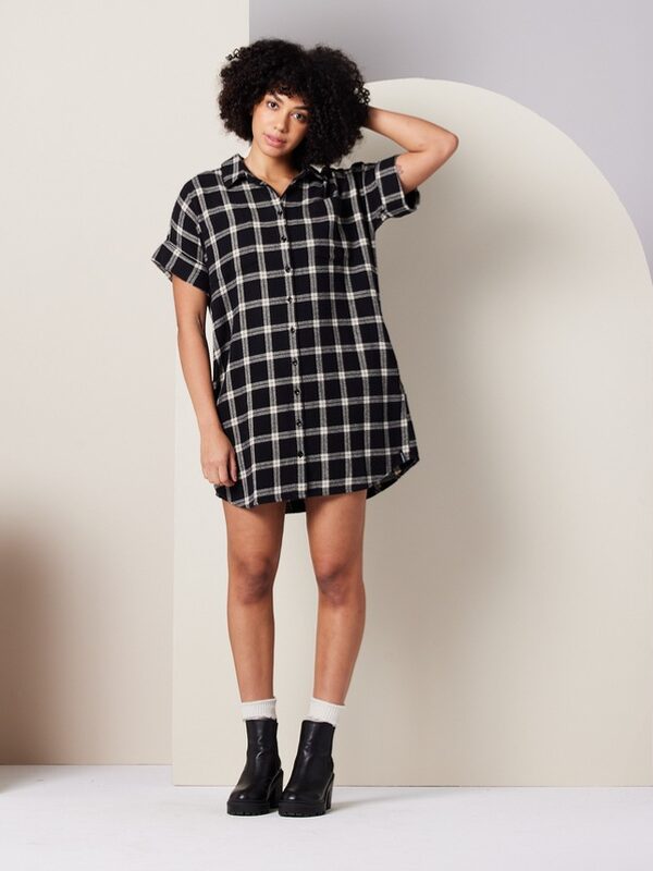 A woman in a checkered dress and black boots stands confidently with one hand on her hip, set against a minimalist background with geometric shapes.