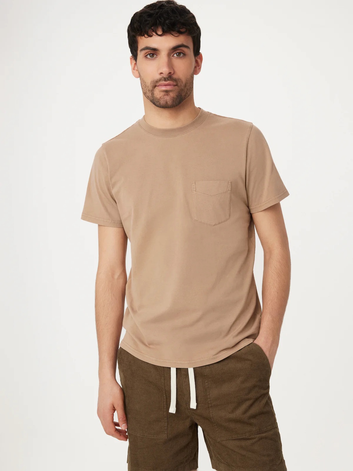 A young man wearing a beige t-shirt and brown trousers standing against a white background.