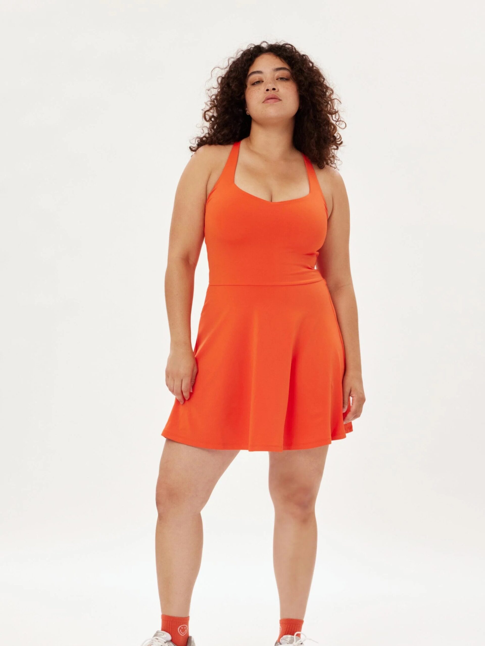 Woman in a bright orange tennis dress and white sneakers standing confidently against a plain background.