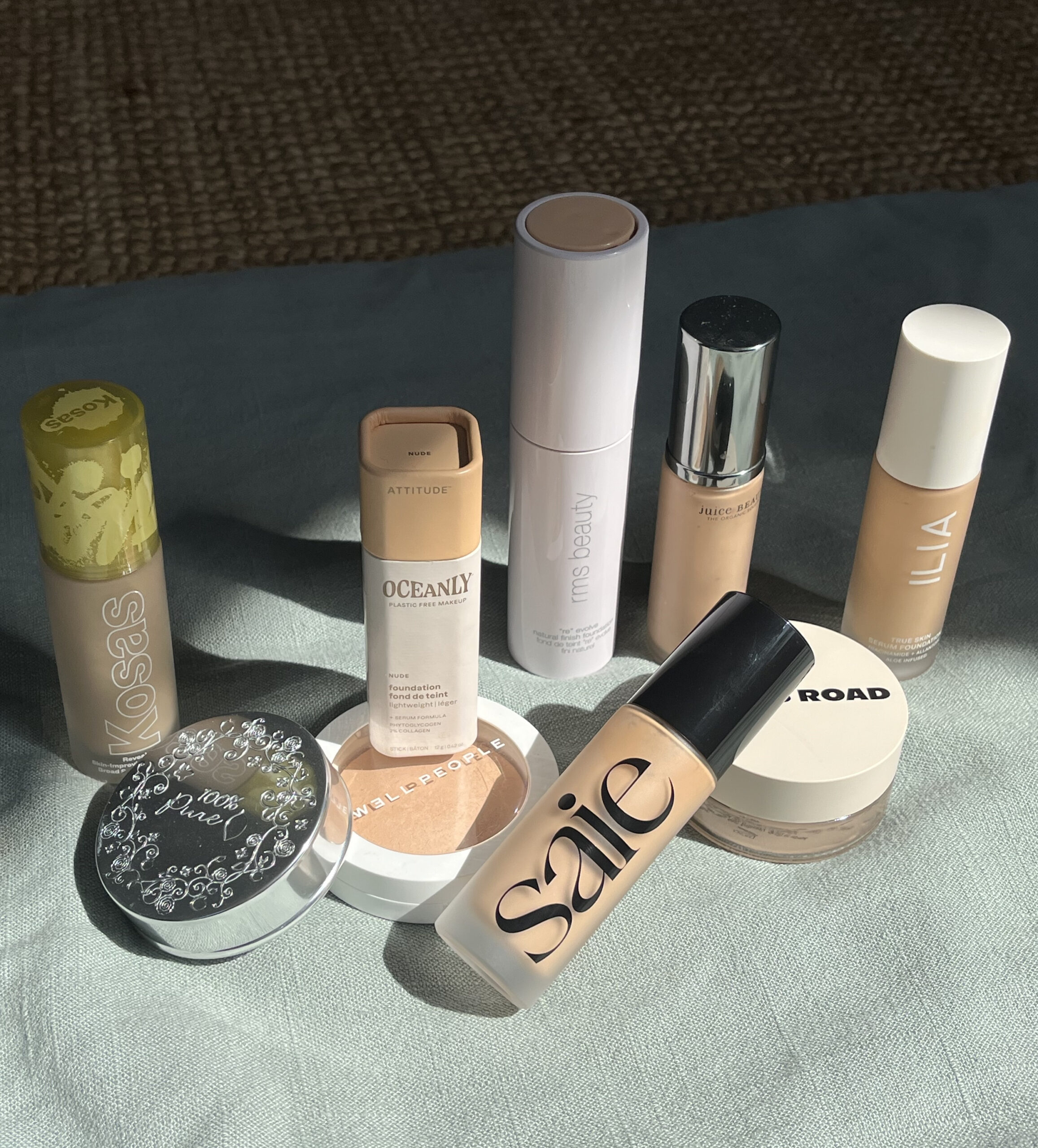 Various brands of makeup products, including foundations and concealers, arranged on a sunlit surface.