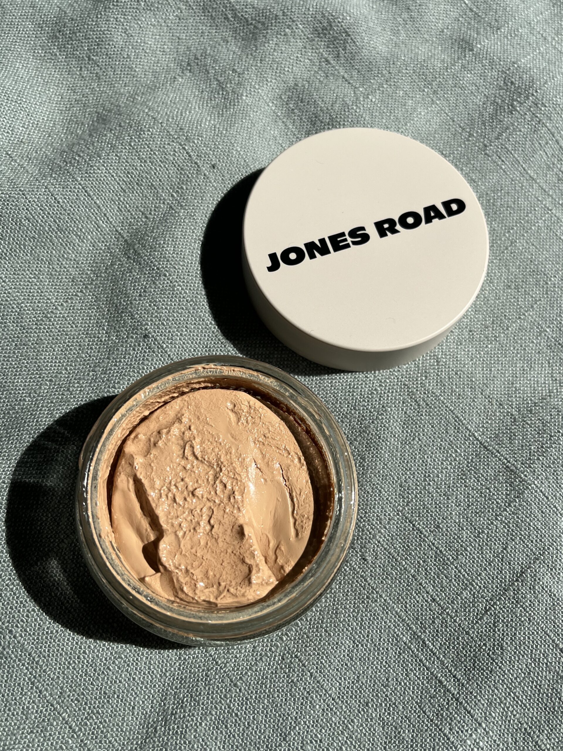 Open jar of jones road cream makeup beside its white lid on a textured blue surface.