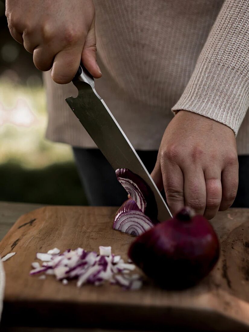 A person is slicing a red onion on a wooden cutting board outdoors, holding the onion with one hand and a knife with the other.