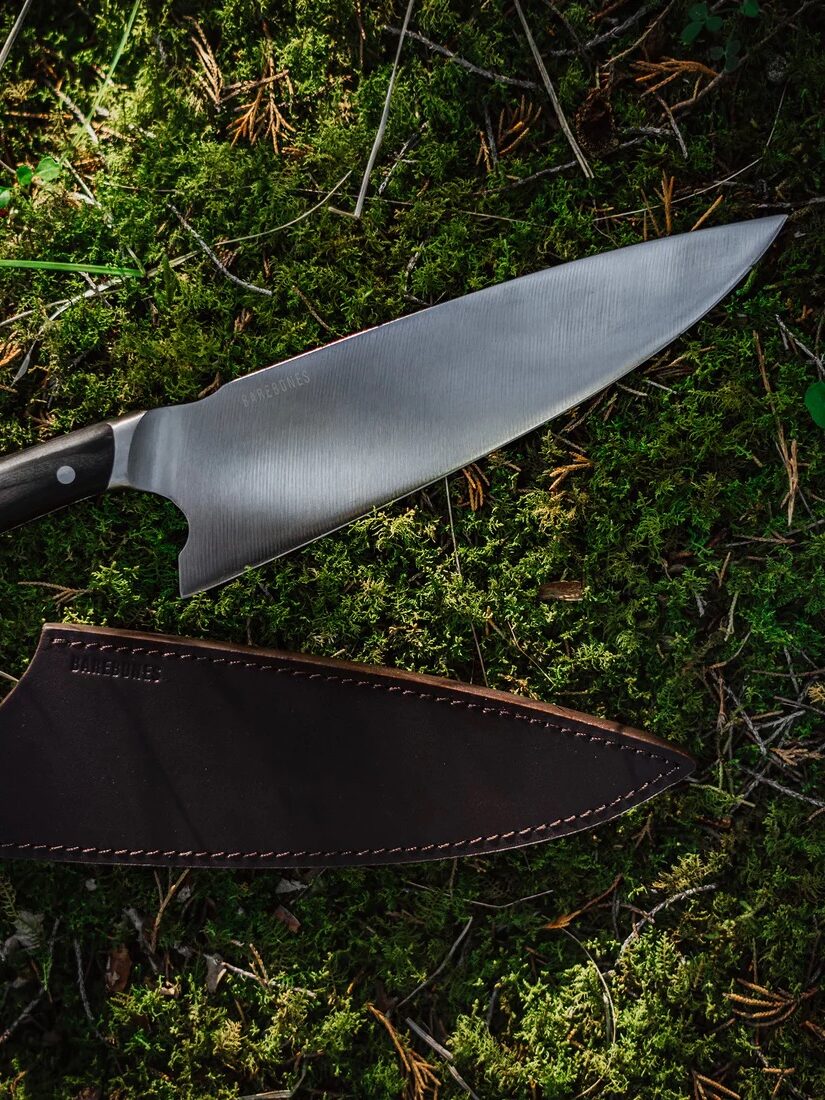 A large chef's knife next to its brown leather sheath, resting on a forest floor covered with pine needles and twigs.