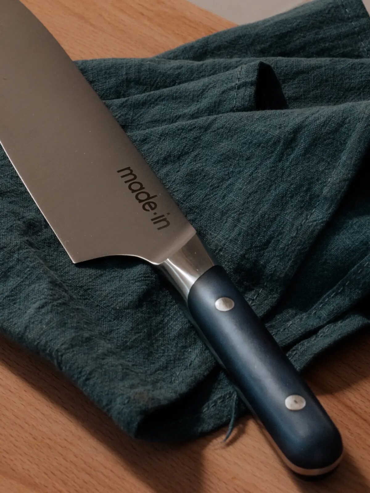A chef's knife with a black handle on a dark green cloth, with the brand "made in" visible on the blade.