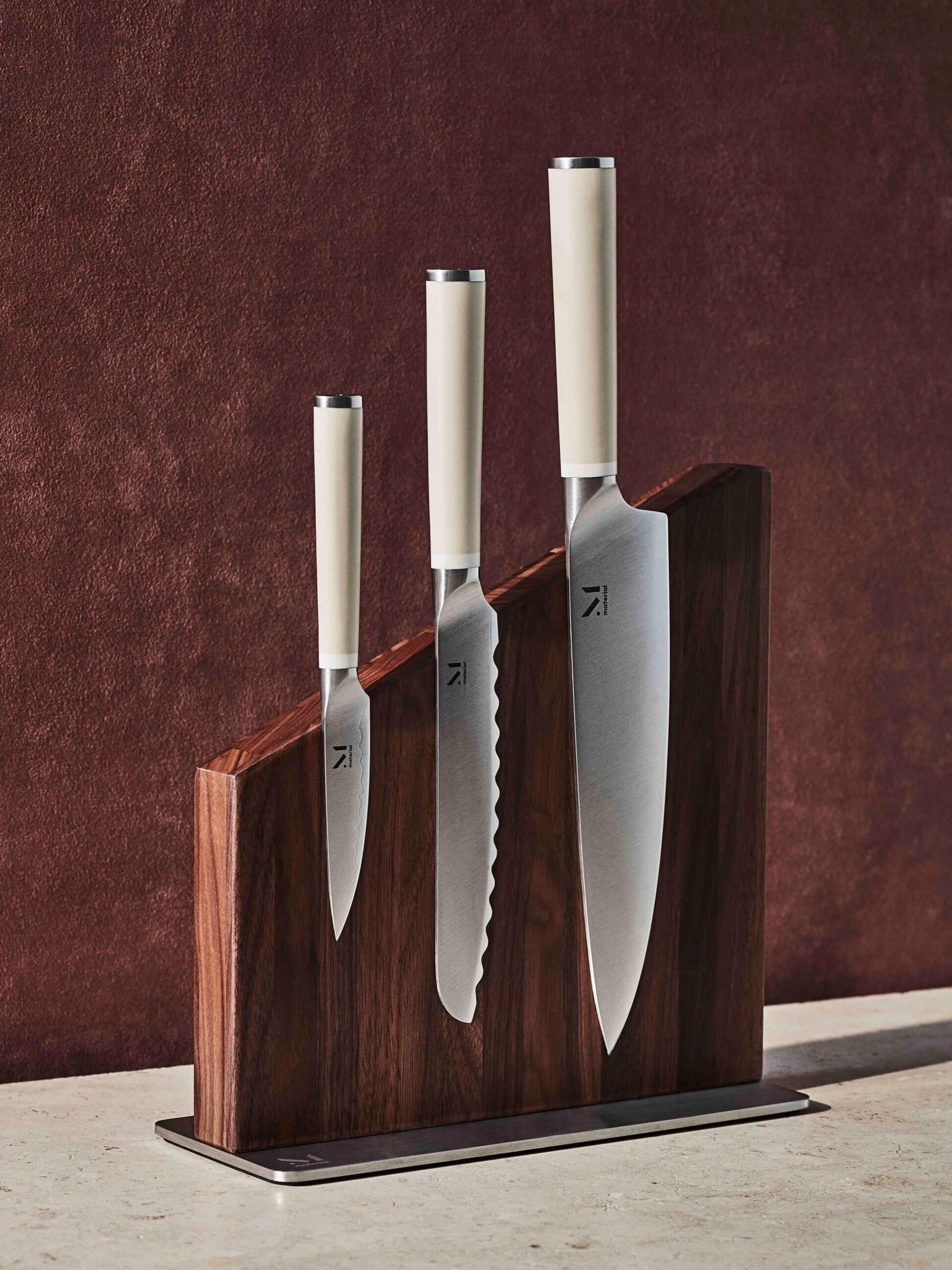 A set of three stainless steel kitchen knives with white handles, displayed upright in a dark wooden block on a textured brown background.
