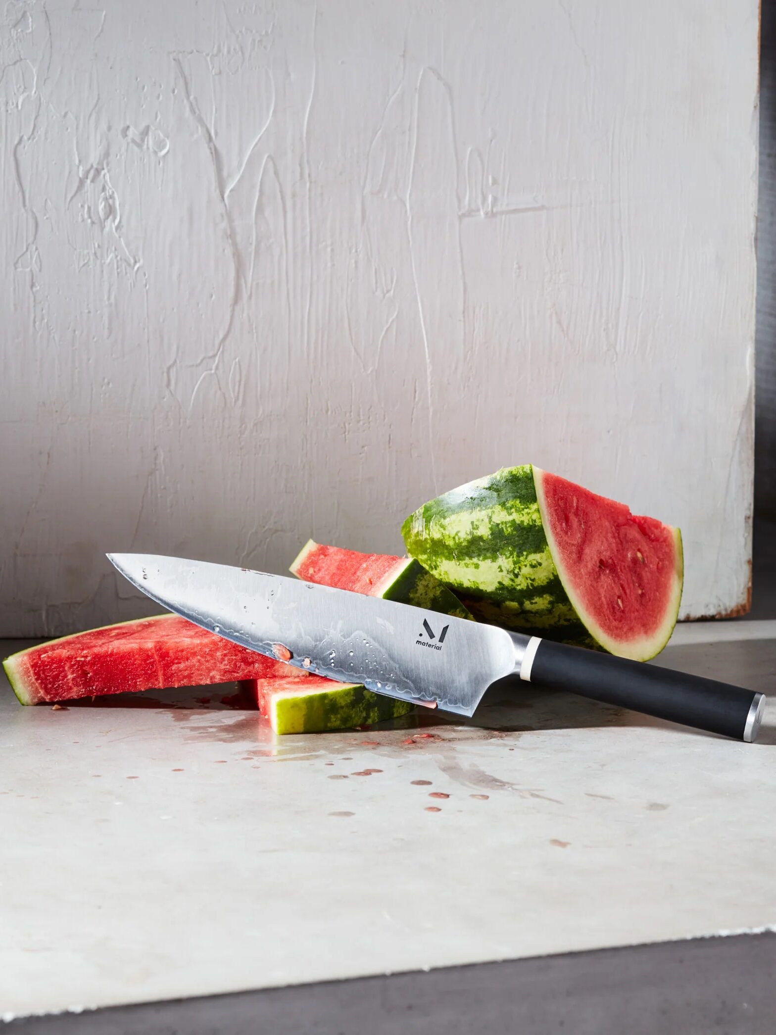 Kitchen knife and freshly cut slices of watermelon on a concrete surface, with light casting shadows on the background wall.