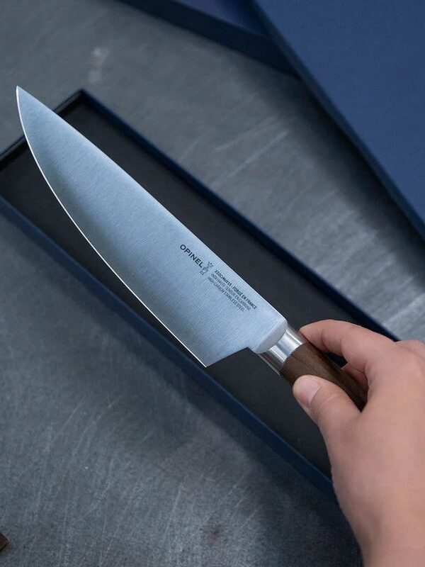 A hand holds a sharp chef's knife with a wooden handle above its box on a grey surface.