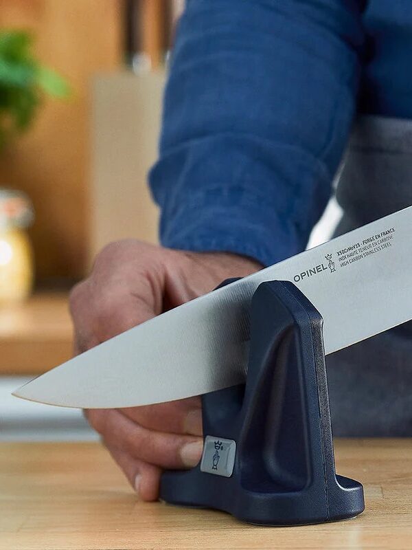 A person in a blue apron sharpens a kitchen knife on a gray sharpening tool on a wooden countertop.
