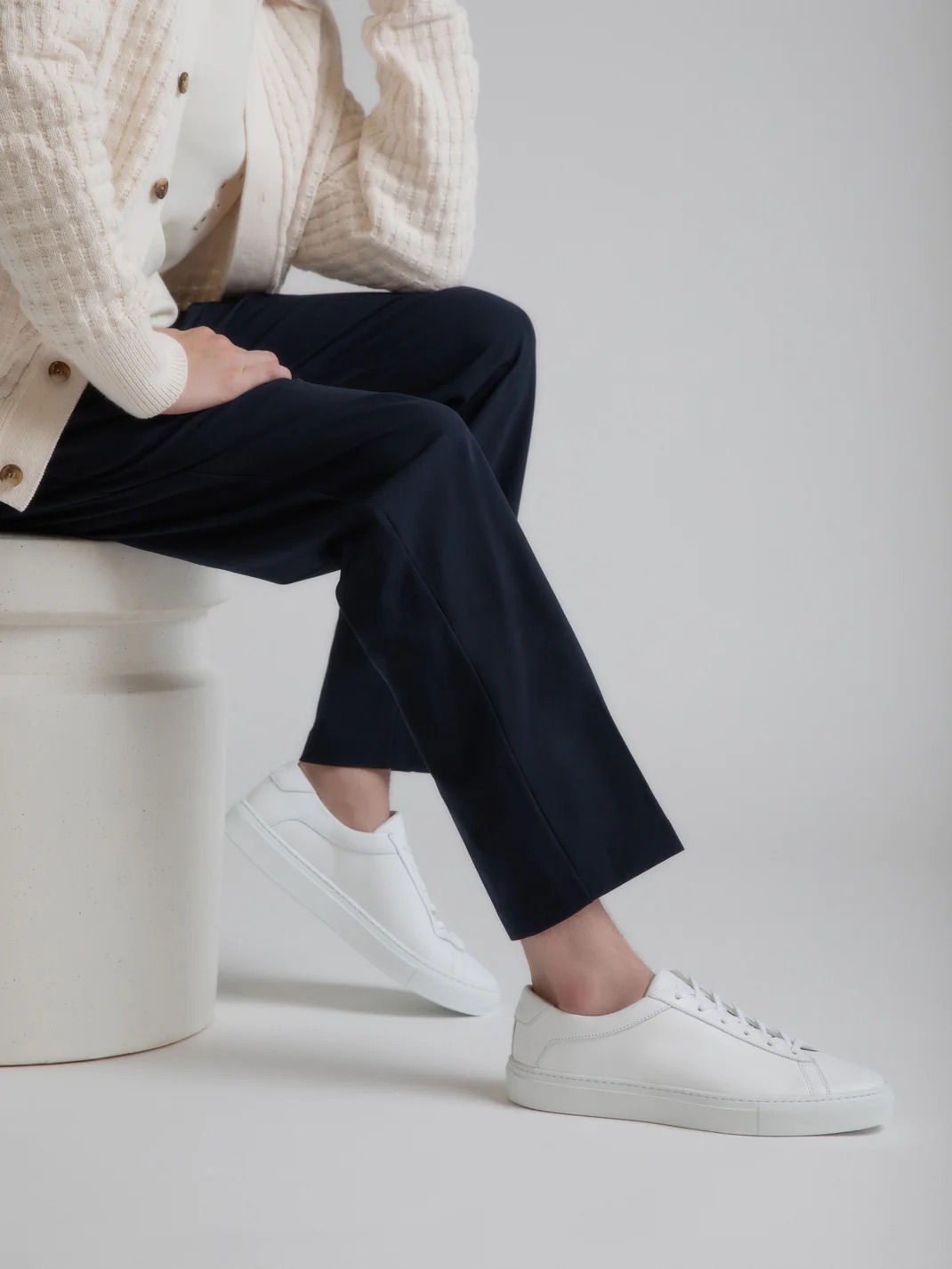 A person seated on a white stool wearing a cream jacket, navy trousers, and white sneakers. only the lower half of the body is visible.