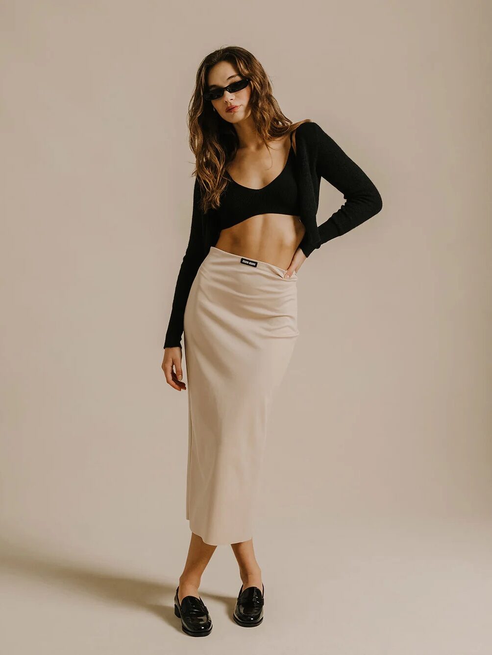 Woman in stylish attire posing confidently, wearing a black crop top, beige skirt, sunglasses, and black loafers against a neutral backdrop.