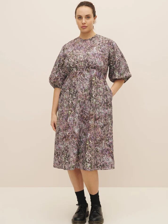A woman stands wearing a floral midi dress with puff sleeves, paired with black ankle boots, against a light beige background.
