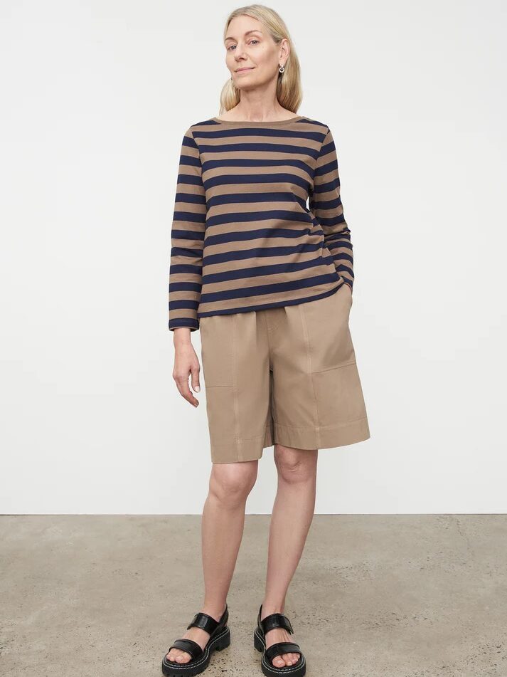 Woman standing in a studio, wearing a striped long-sleeve top and khaki shorts, paired with black sandals.