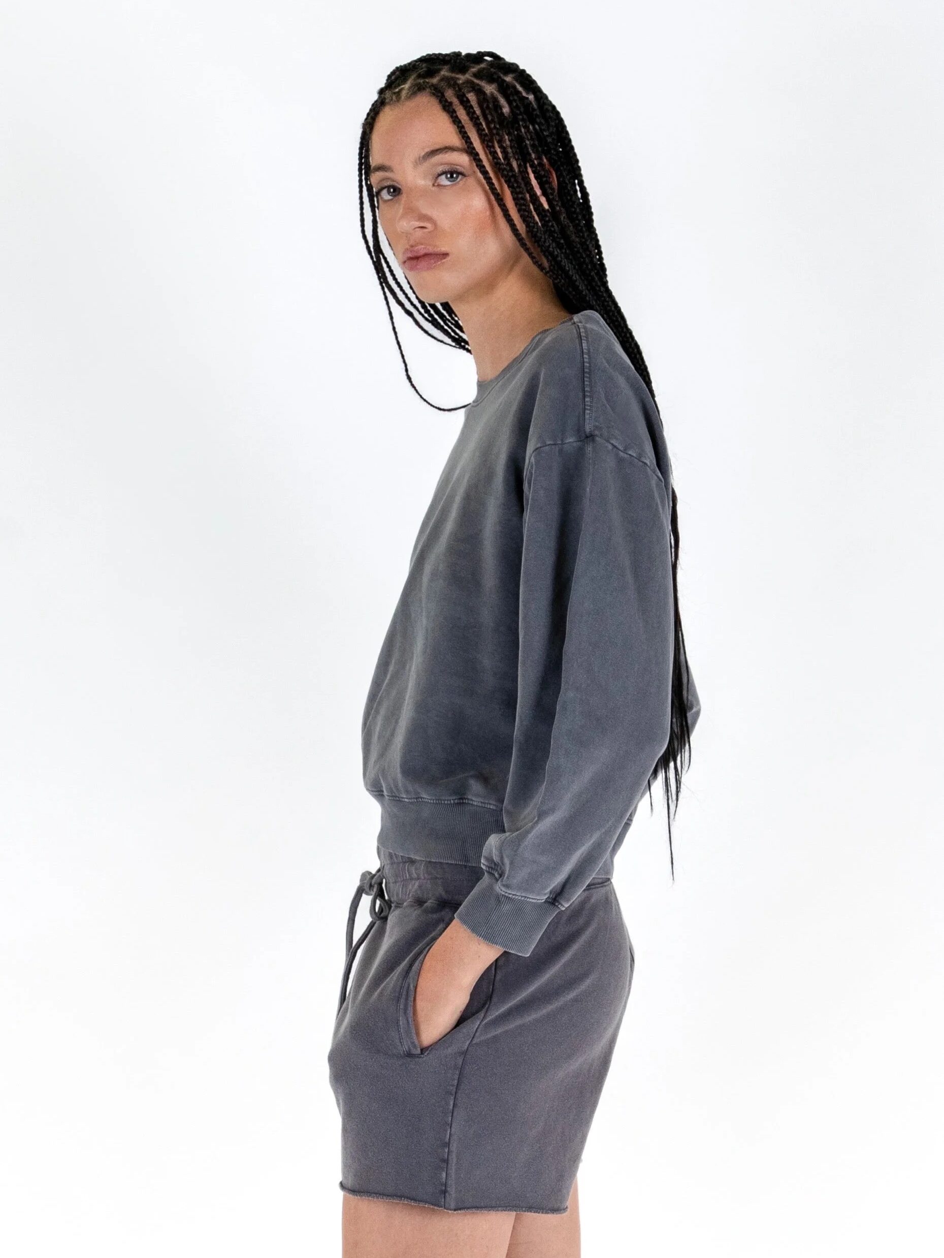 Woman in a gray sweatshirt and skirt looking over her shoulder with wet hair styled in thin braids against a white background.