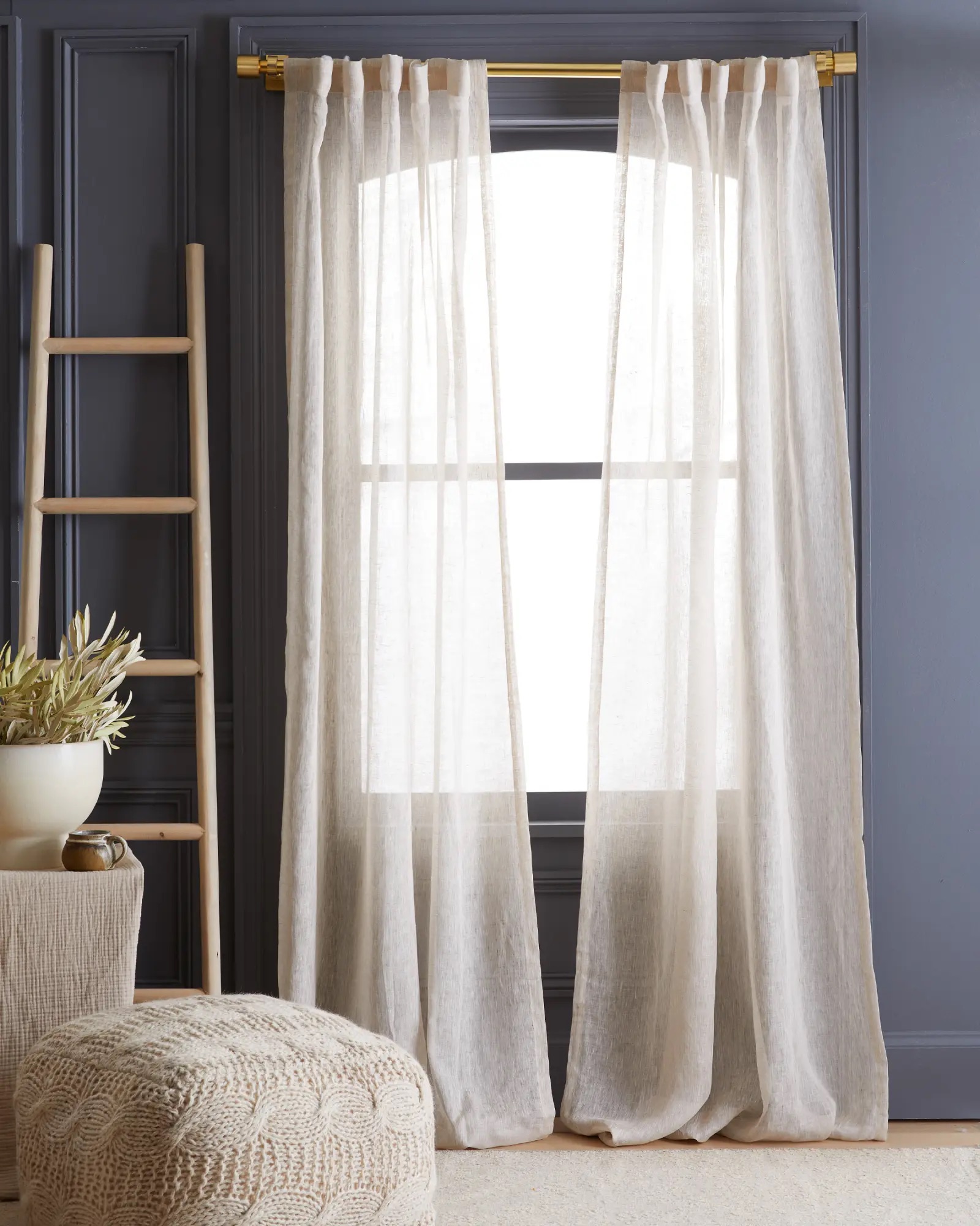 Sheer white curtains hanging on a gold curtain rod in front of a window, with a grey wall, a wooden ladder, and a beige ottoman.