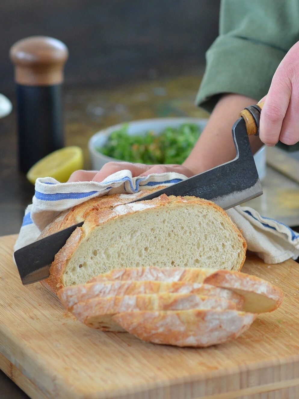 A person cutting a loaf of bread on a wooden cutting board, with a bowl of greens and another person in the background.