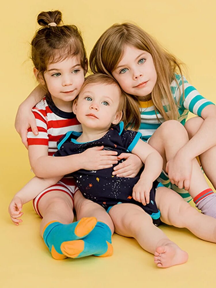 Three young children posing together on a yellow background, two girls hugging a baby boy wearing colorful clothes.