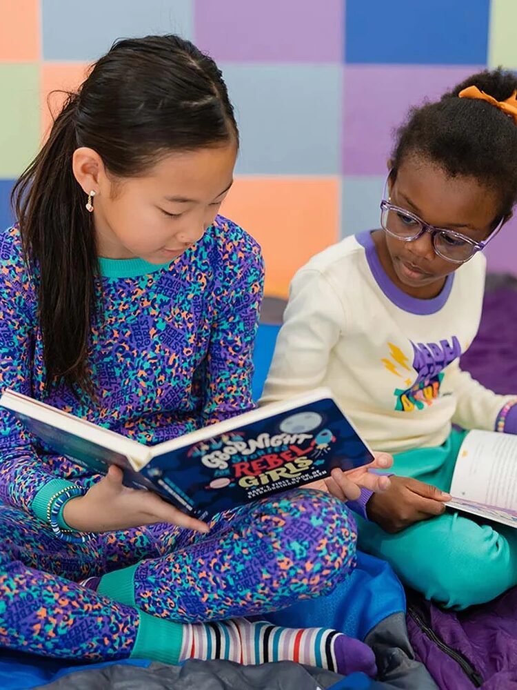 Two young girls reading a book together on a colorful mat, one asian and the other black, both smiling and engaged in the story.