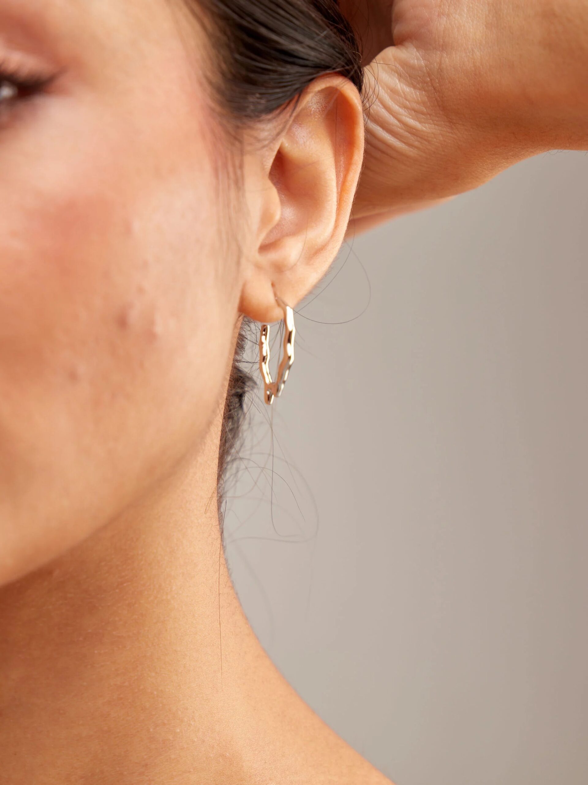 Close-up of a woman's ear wearing a delicate, gold hoop earring, with her hand gently lifting her hair.