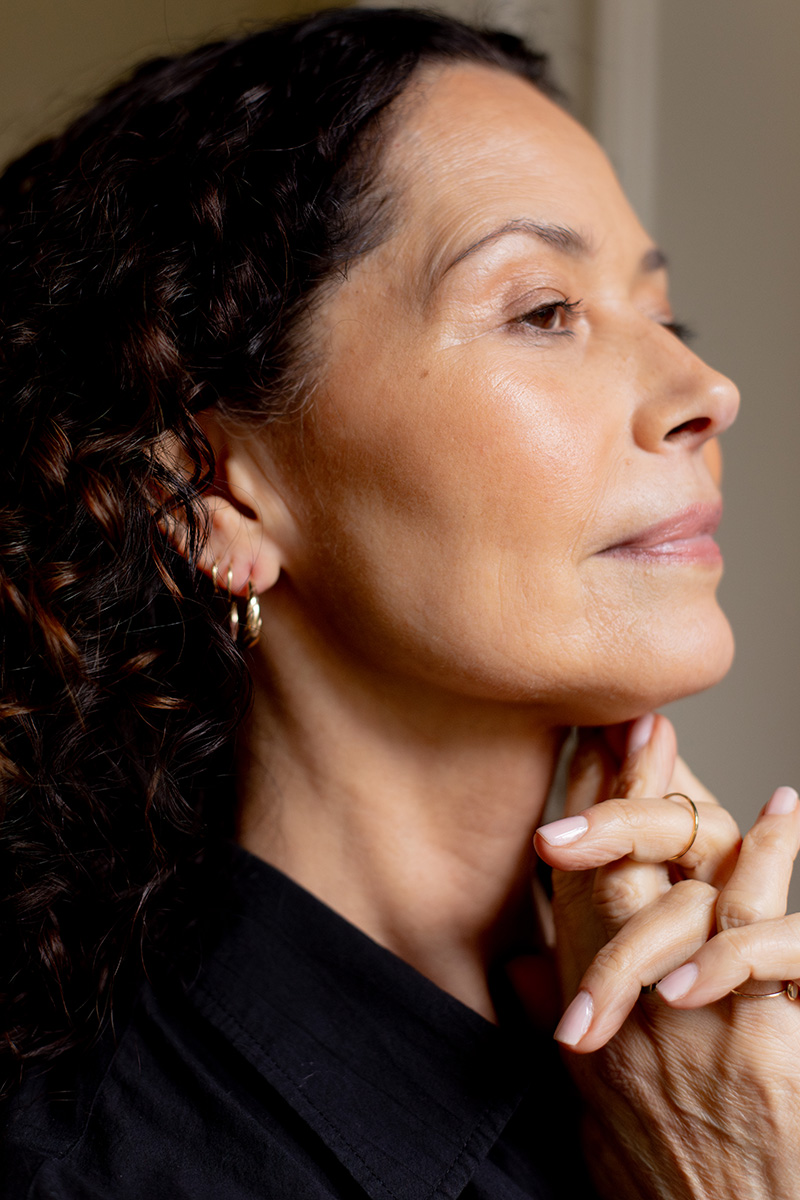 Profile view of a woman with curly hair, touching her face gently, wearing earrings and a black shirt, with a contemplative expression.