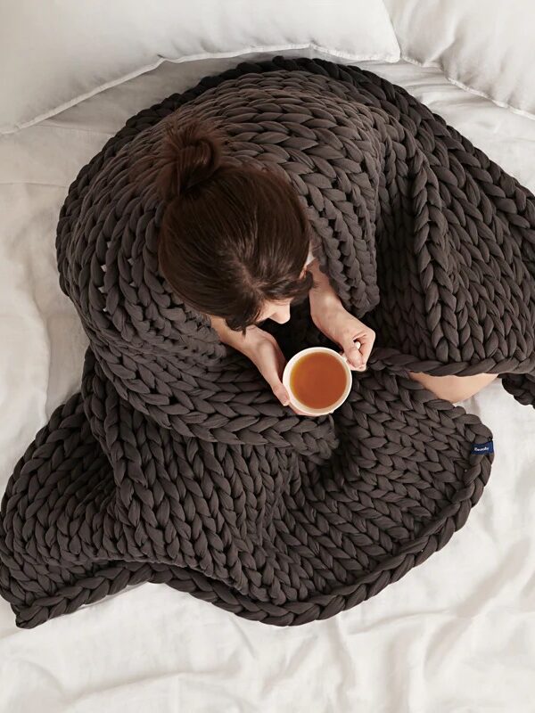 Aerial view of a person bundled in a chunky knit blanket, holding a mug, sitting on a bed.
