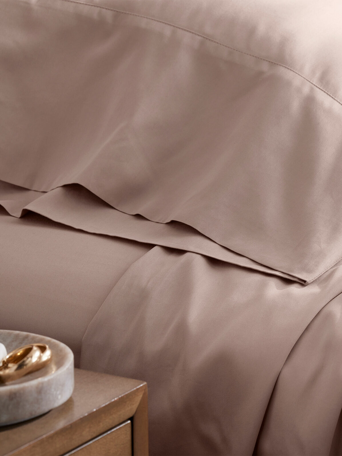 Luxurious beige cotton bedding with soft wrinkles on a cozy bed next to a wooden bedside table featuring a decorative item.