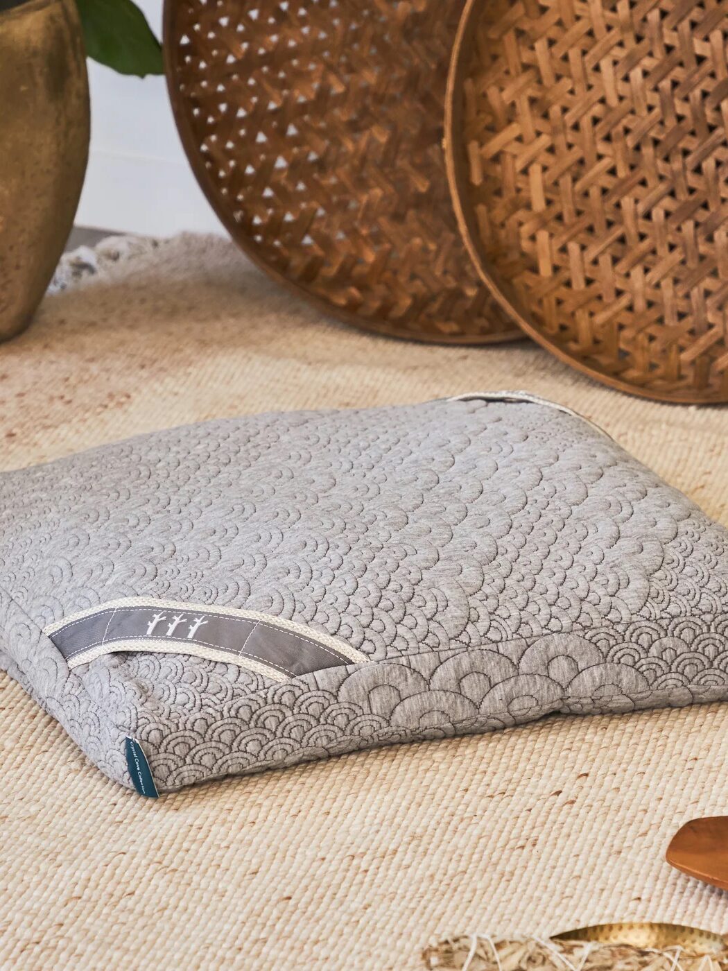 A gray meditation cushion with a white pattern, placed on a woven mat, surrounded by potted plants and wooden artifacts.