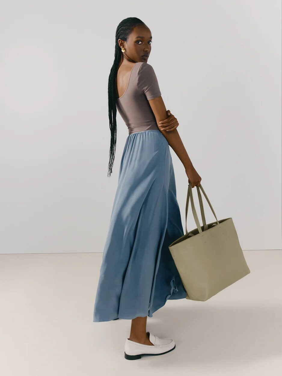 A woman with braided hair, wearing a lavender top and blue skirt, walks looking over her shoulder while holding a beige tote bag.