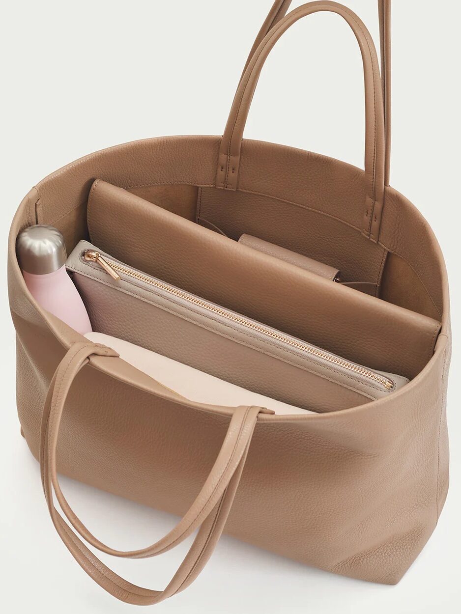 An open brown leather tote bag displaying internal compartments with a pink wallet and a stainless steel water bottle inside.