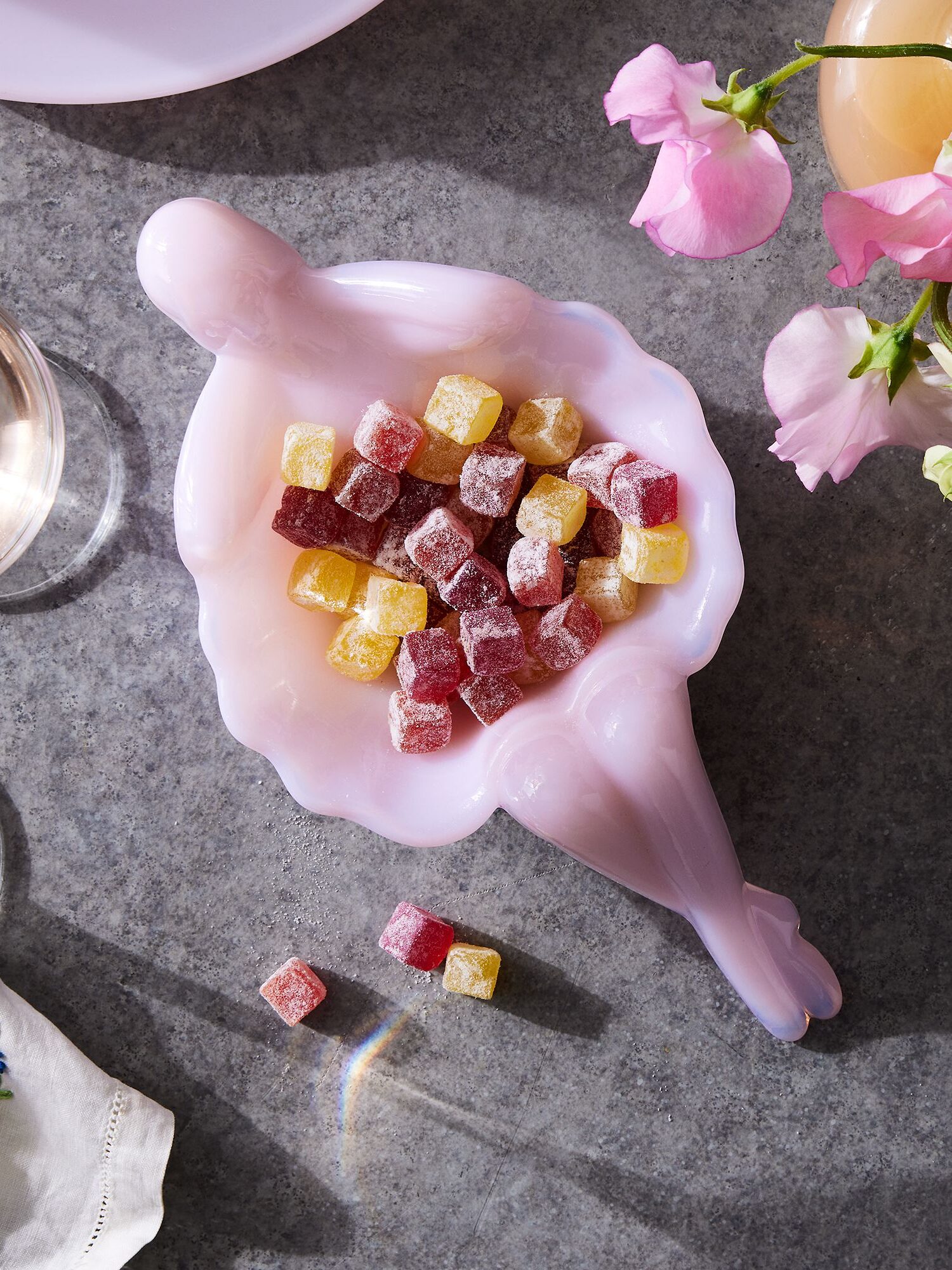 A pink glass shell-shaped dish filled with colorful sugared fruit jelly cubes, accompanied by pink drinks and floral décor on a grey surface.