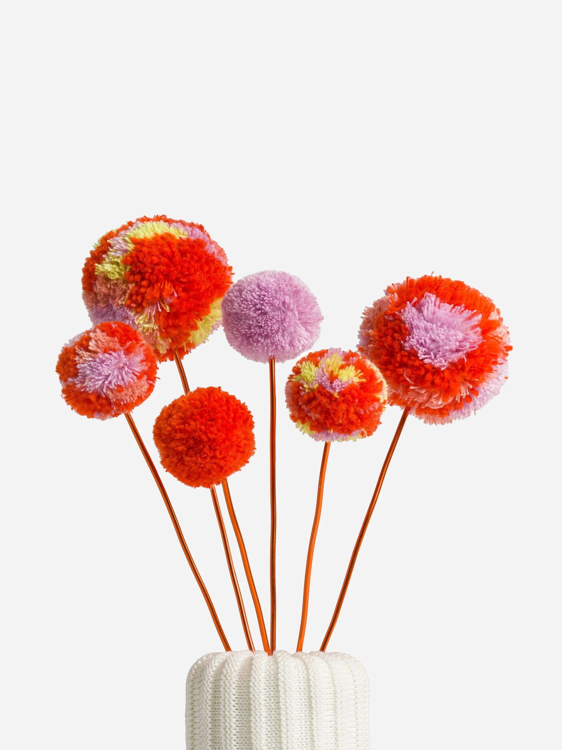 A collection of colorful pompom flowers on thin stems, arranged in a white vase against a plain background.