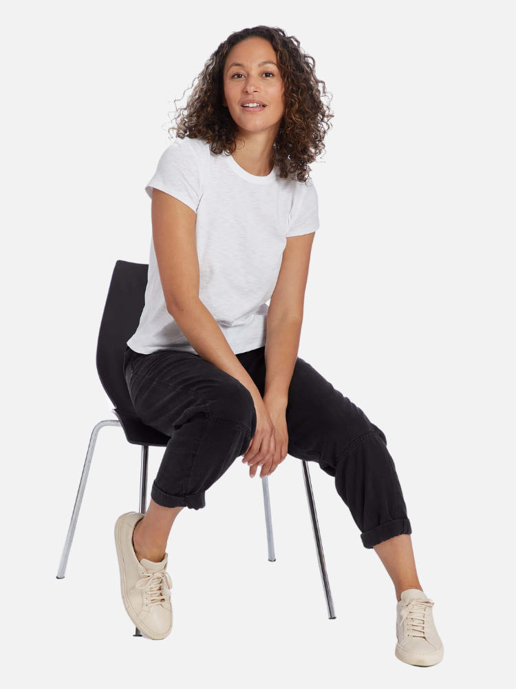 A woman sitting on a chair wearing a white t-shirt and black pants, smiling at the camera, with a white background.