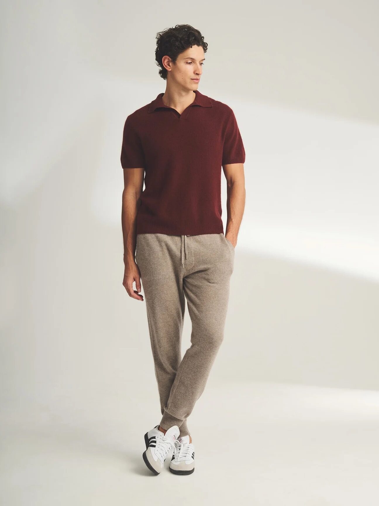 A man stands in a studio, wearing a burgundy polo shirt, grey trousers, and white sneakers, looking to his right with a neutral expression.
