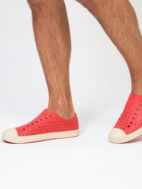 A person wearing red perforated slip-on shoes stands against a white background, showing only their lower legs and feet.