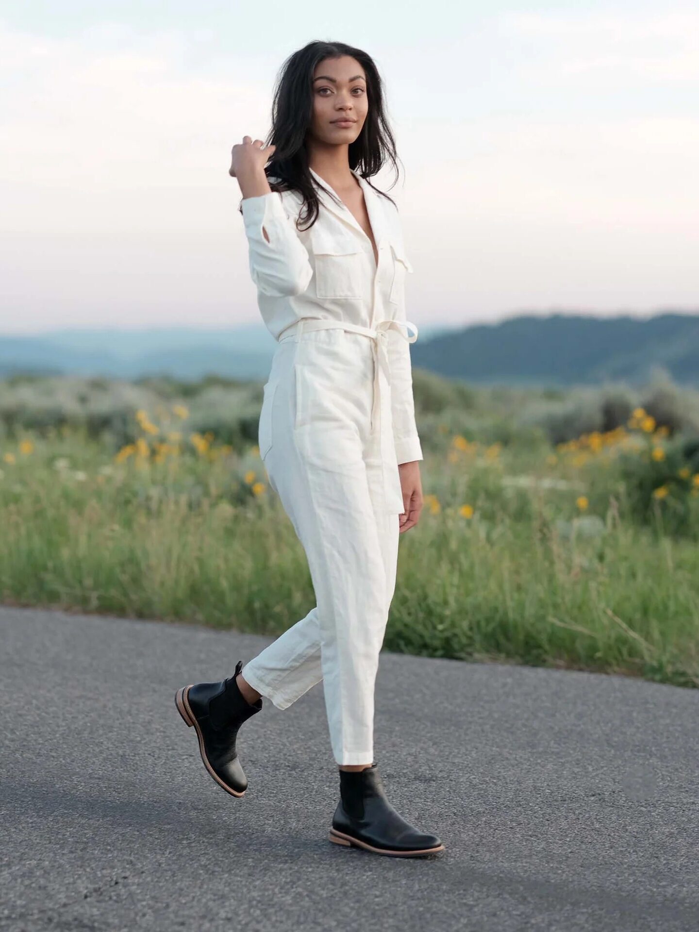 A woman in a white jumpsuit and black boots walks along a rural road with fields of yellow flowers in the background at dusk.