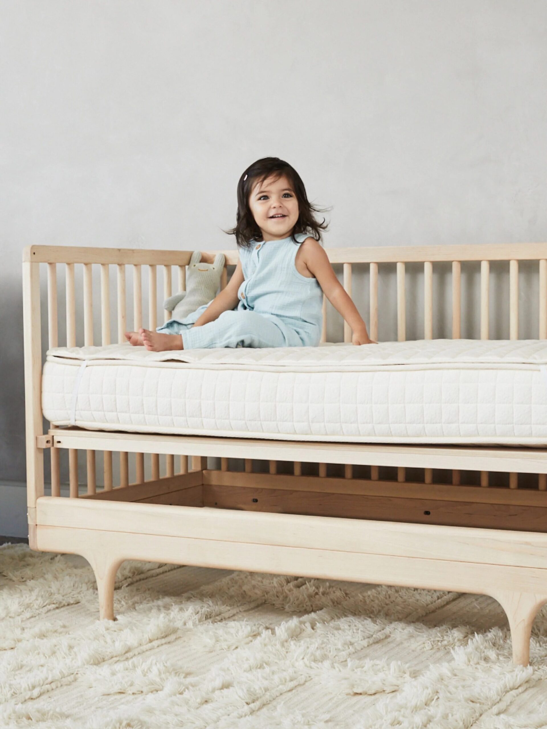 A smiling child sits on a wooden toddler bed in a neutrally decorated room.