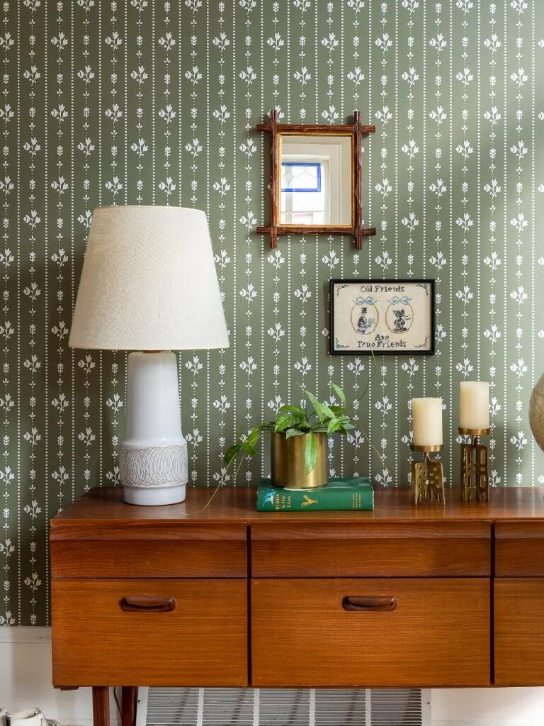 A vintage wooden sideboard with a lamp, plant, globe, and candles against a floral wallpaper background.