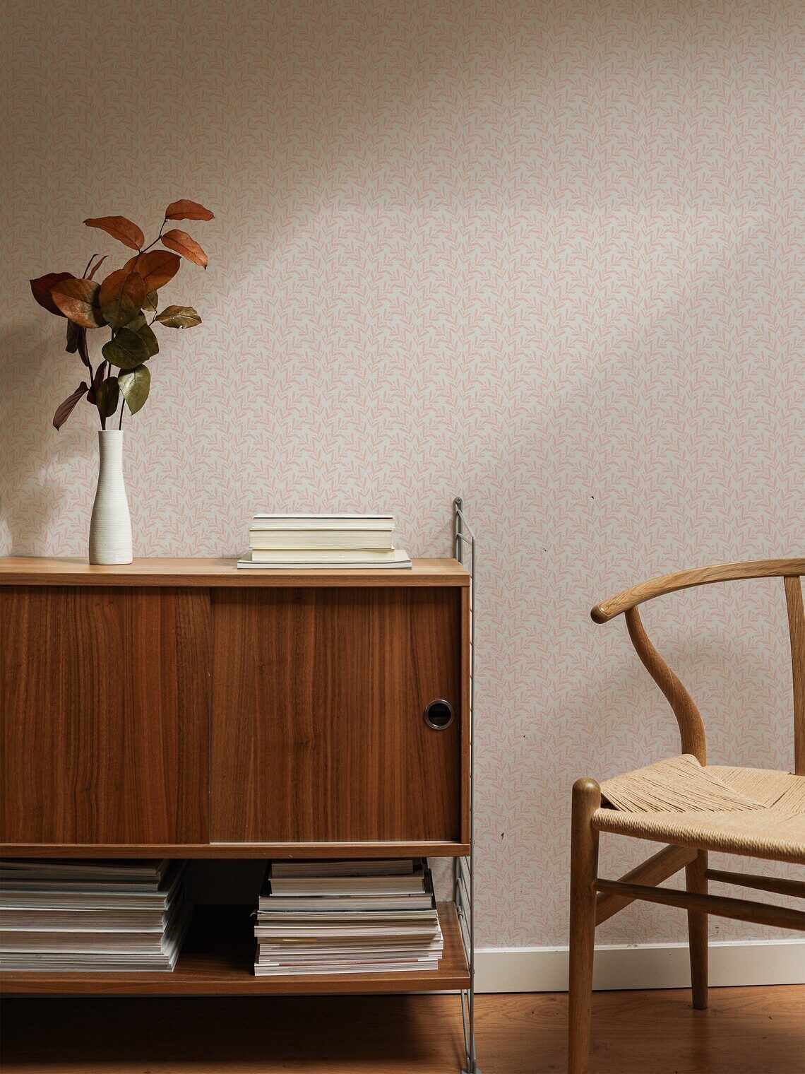 A cozy room corner featuring a wooden sideboard and chair against a wallpapered wall, with a vase of leaves and books on top of the sideboard.