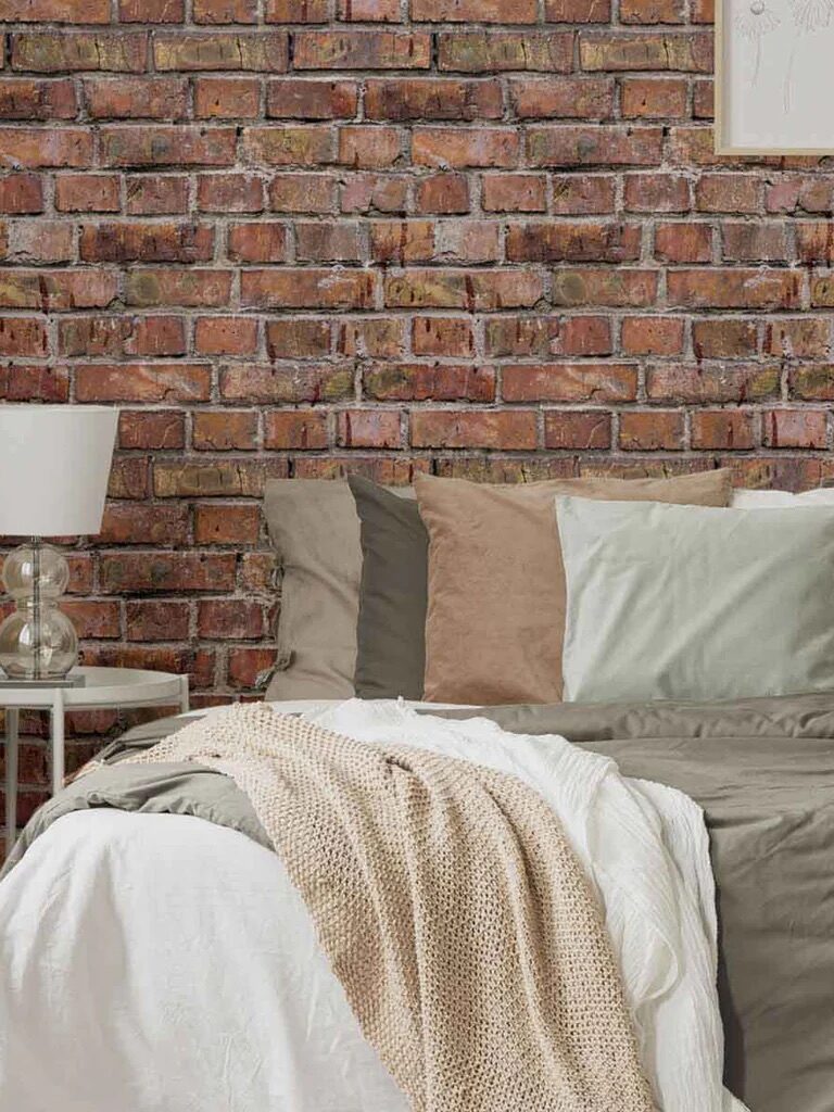 A cozy corner with a bed covered in a beige throw blanket, assorted pillows, and a lamp on a small side table against a brick wall background.