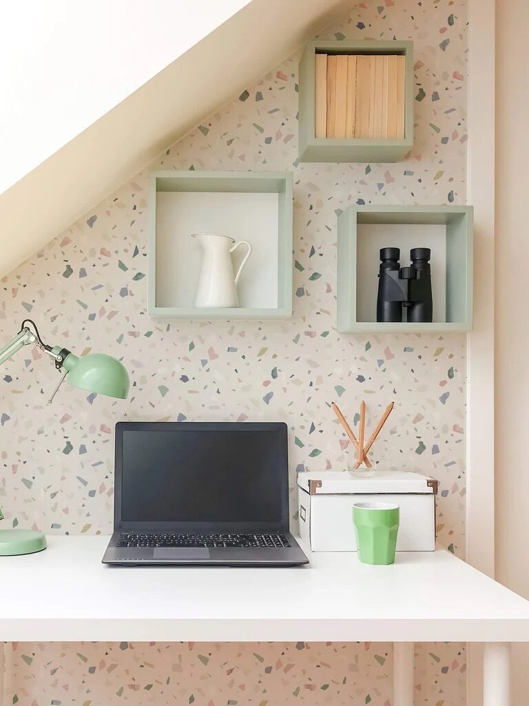 A neat home office nook with a laptop on a white desk, green lamp, and decorative shelves against a patterned wallpaper.